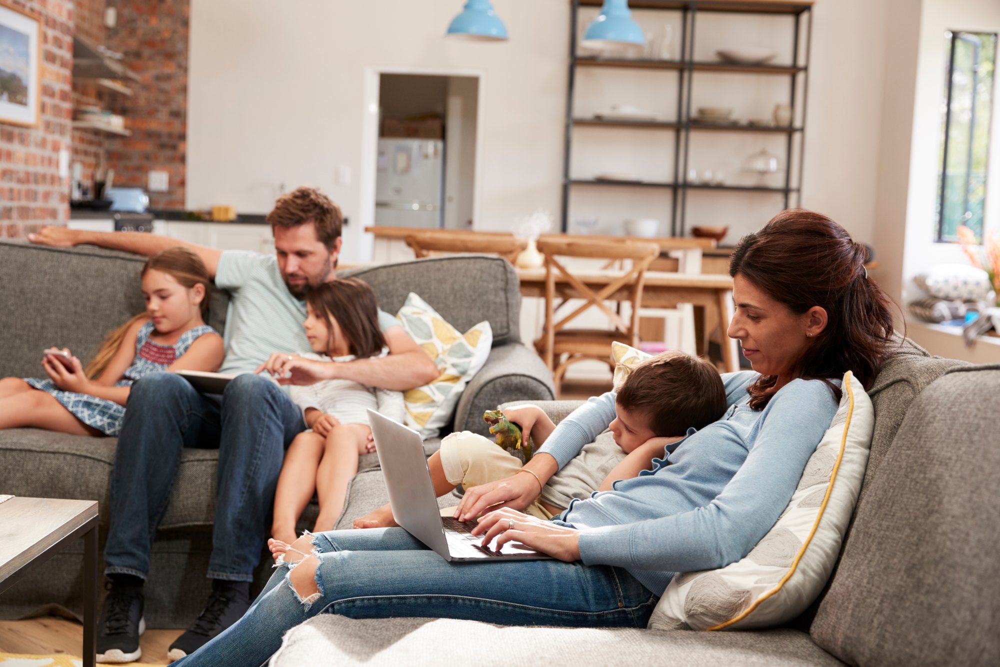 Family Sit On Sofa In Open Plan Lounge Using Technology